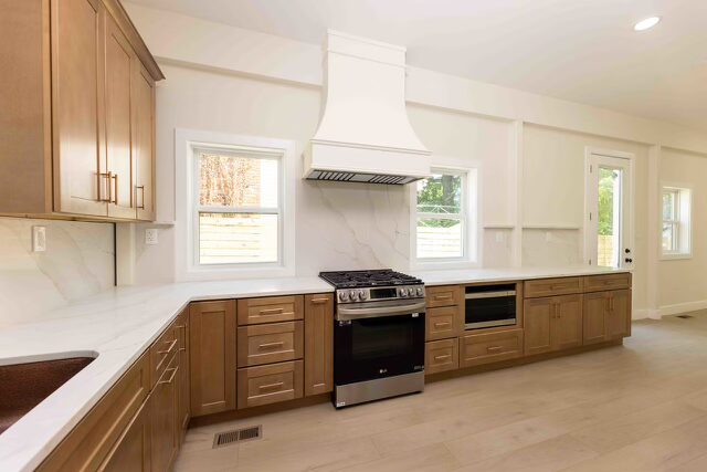 Custom wood range hood above a modern gas stove in a kitchen with light-colored cabinetry and marble countertops.